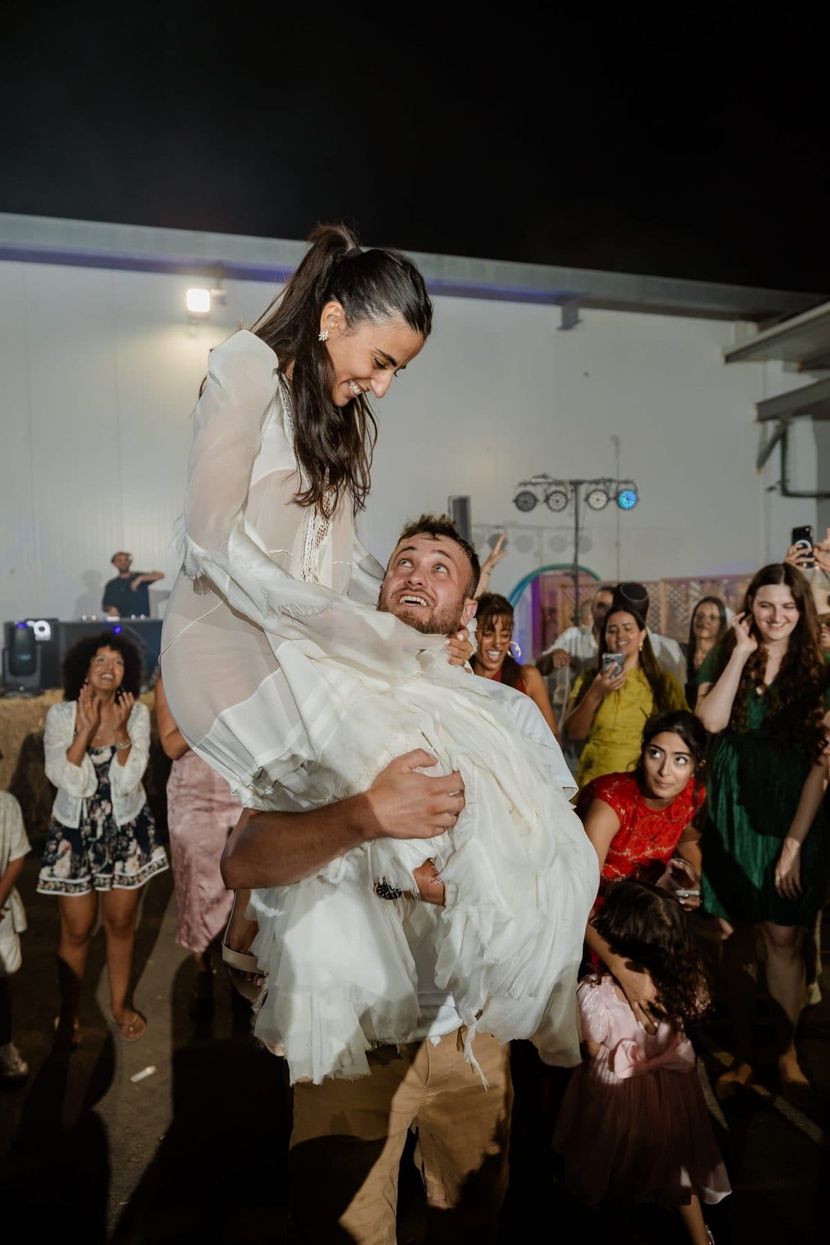 החתן מרים את הכלה בריקוד, והאורחים צוהלים סביבם